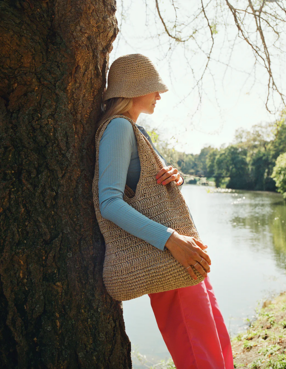 Beige raffia bucket-hatt på modell i profil ved tre og elv, sommerlig setting