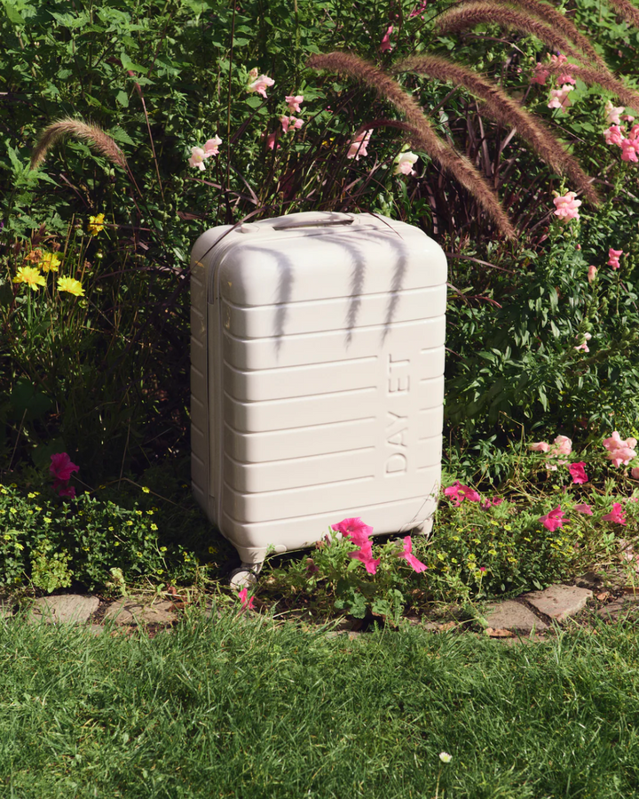 Pastel beige hardskall kabinkoffert med preget logo, frontsett i hageomgivelser med blomster