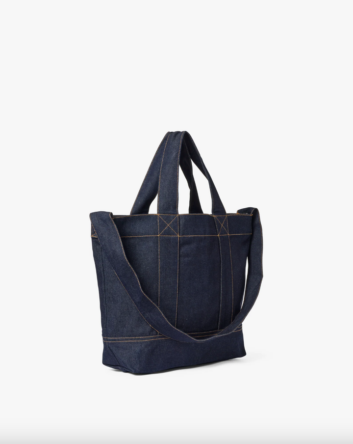 Mørk denim tote-veske med crossbody-rem og kontrastsømmer, sidevinkel mot hvit bakgrunn