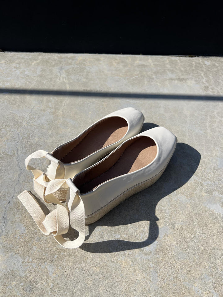 Hvite ballerinasko med ankelstropper i skinn og jute-såle, flat lay ovenfra på betongfliser