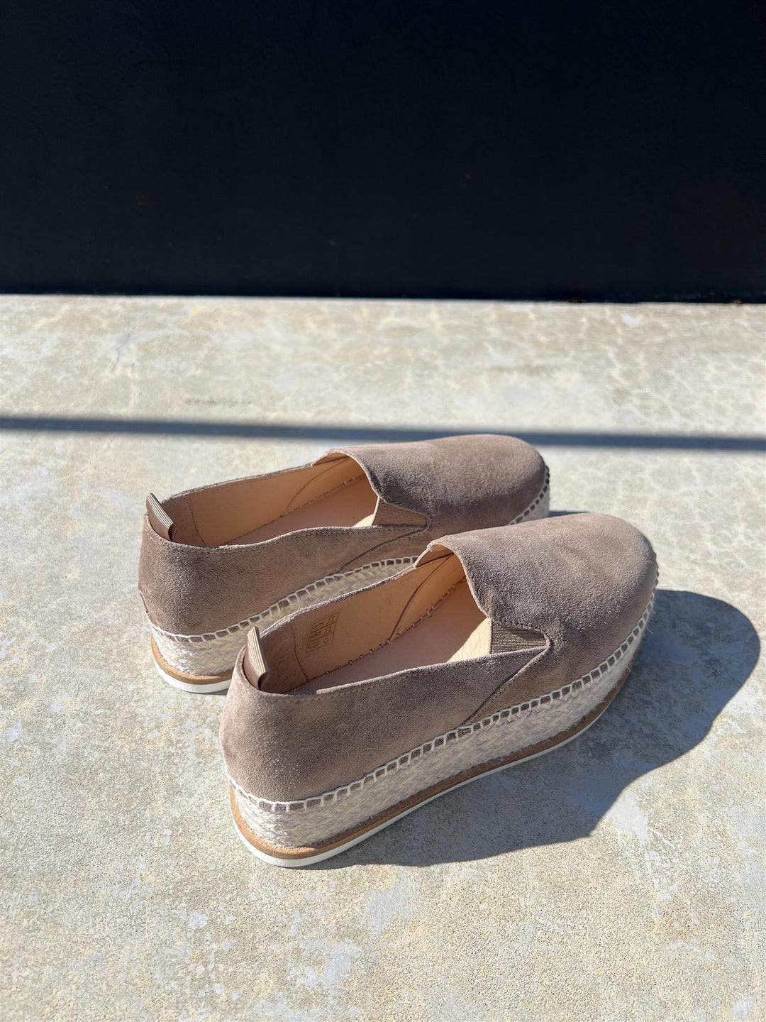 Taupe espadrillesko med jute-såle fotografert ovenfra på steinhelle i sollys, viser pull-tab