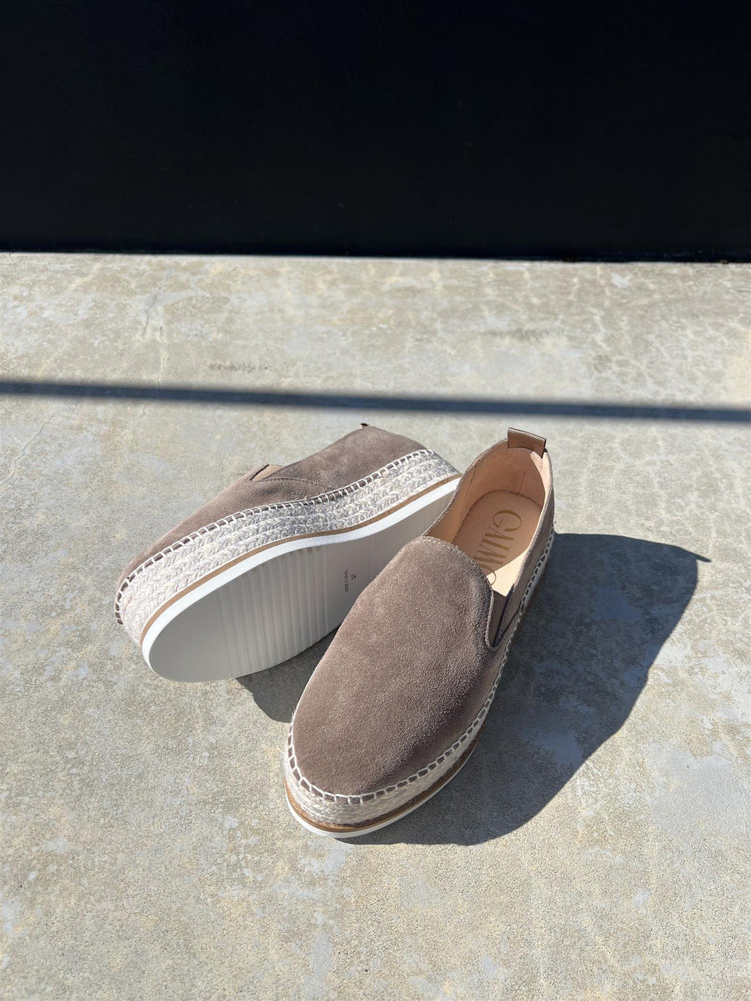 Taupe espadrille-sko med jute-såle og hvit bunn, fotografert ovenfra på steinfliser i sollys