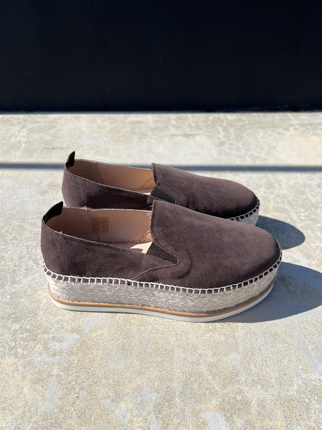 Sidevinkel av chokoladebrun espadrille slip-on sko med platåsåle i jute og gummi på steinfliser