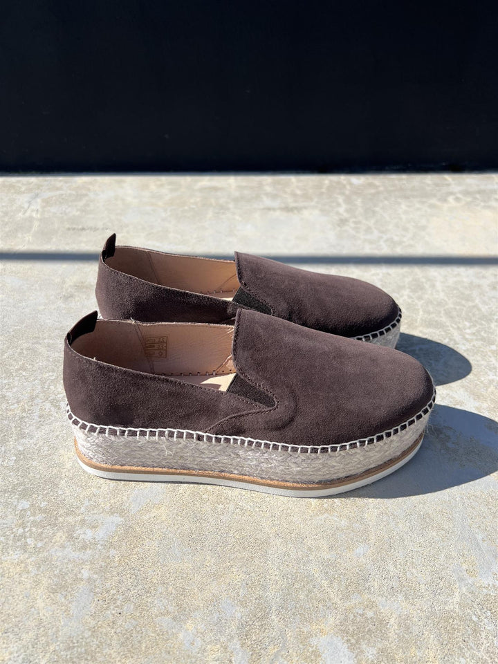 Sidevinkel av chokoladebrun espadrille slip-on sko med platåsåle i jute og gummi på steinfliser