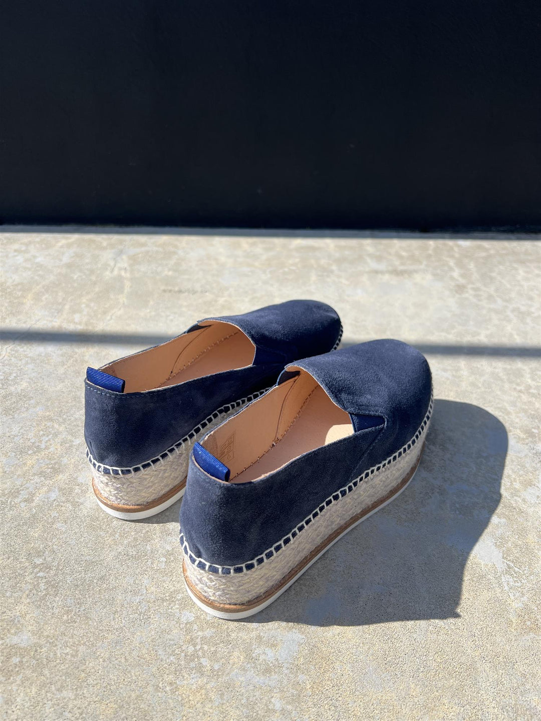 Bakside av mørkeblå espadrilles med jutesåle og hvit gummibunn på steingulv i sollys