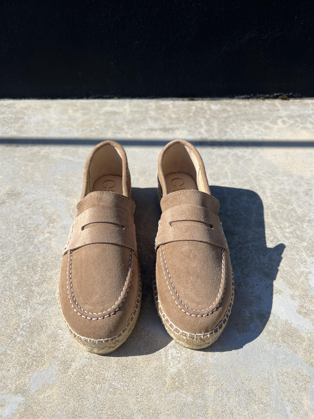 Beige semsket loafers med penny-rem og espadrille-såle, frontsett ovenfra på betongfliser i sollys