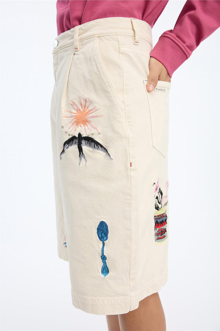 Sidevinkel av beige shorts med broderte motiver inkludert sol, palme og sløyfe på modell