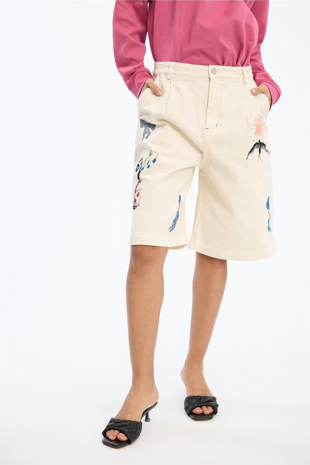 Beige shorts med broderte motiver på modell, frontsett med hender i lommer og rosa genser