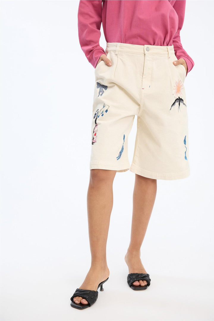 Beige shorts med broderte motiver på modell, frontsett med hender i lommer og rosa genser