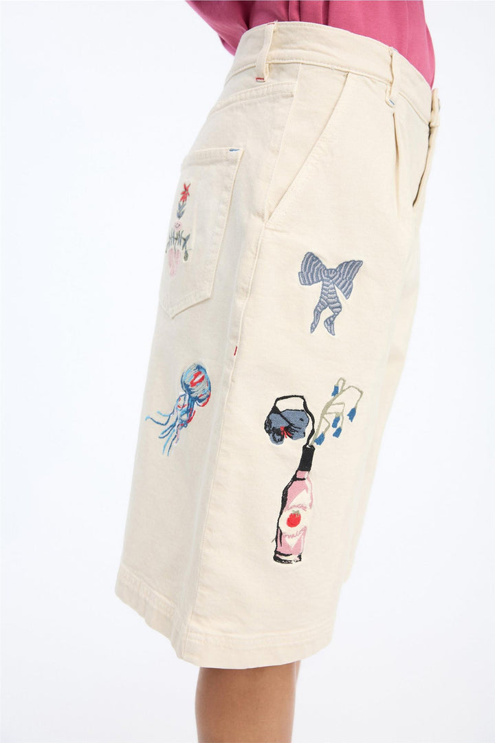 Sidevinkel av beige shorts med broderte motiver inkludert sløyfe, flaske, manet og palme på modell