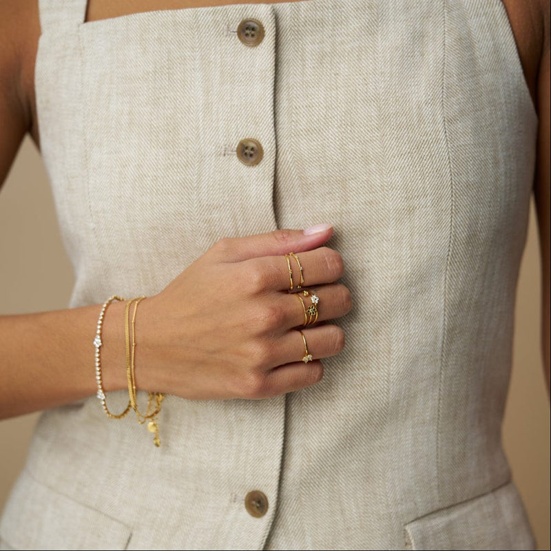 Gulltonet tennis armbånd med blomsterdetaljer og ringer på modellens hånd mot beige vest