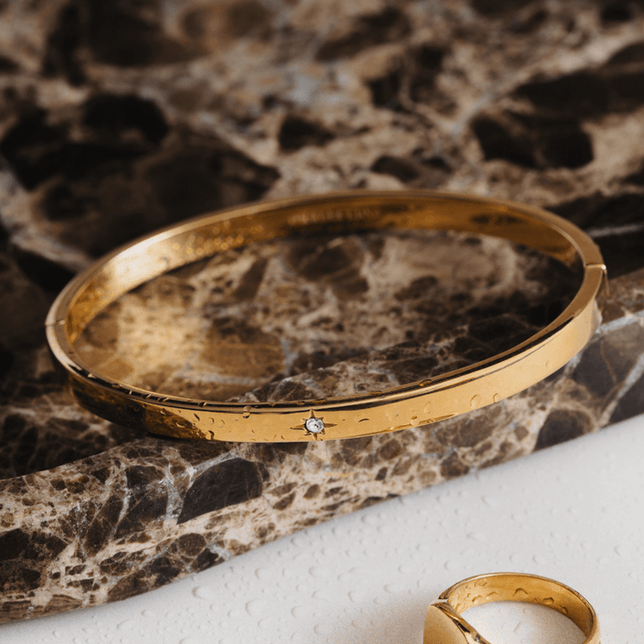 Gulltonet bangle-armbånd med krystalldetalj på marmorstein, flat lay ovenfra med matchende ring